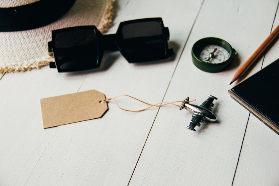 Flat lay of travel essentials including airplane model, sunglasses, compass, and hat on a table.